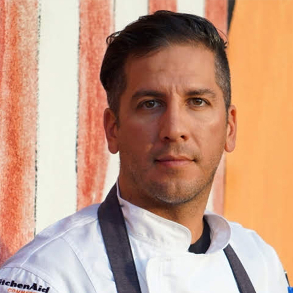 A man wearing a white chef’s coat and dark apron stands in front of a wall with red and orange vertical stripes, looking at the camera.