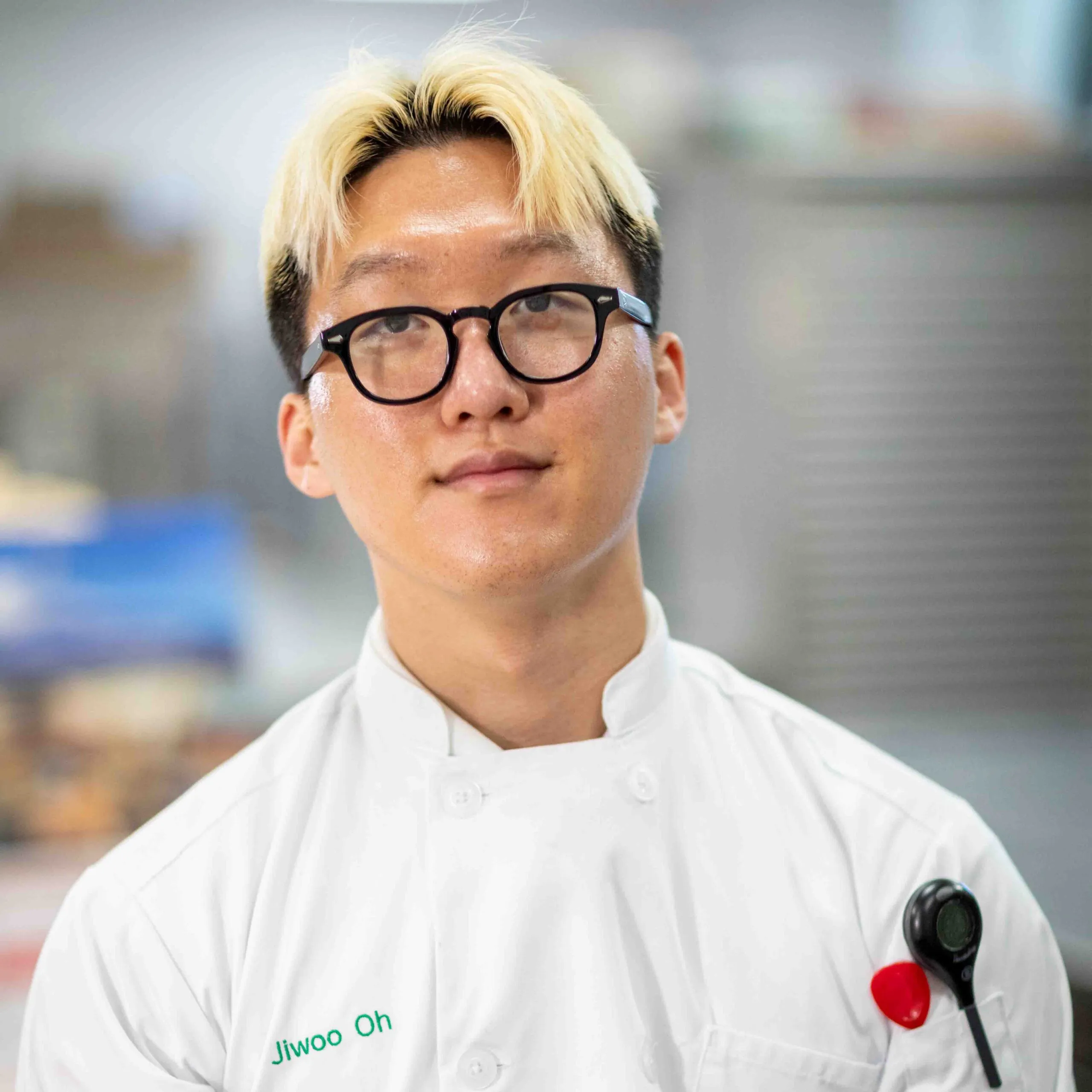 A person with blond hair and glasses wears a white chef's coat with the name "Jiwoo Oh" embroidered on it, standing in a kitchen setting.