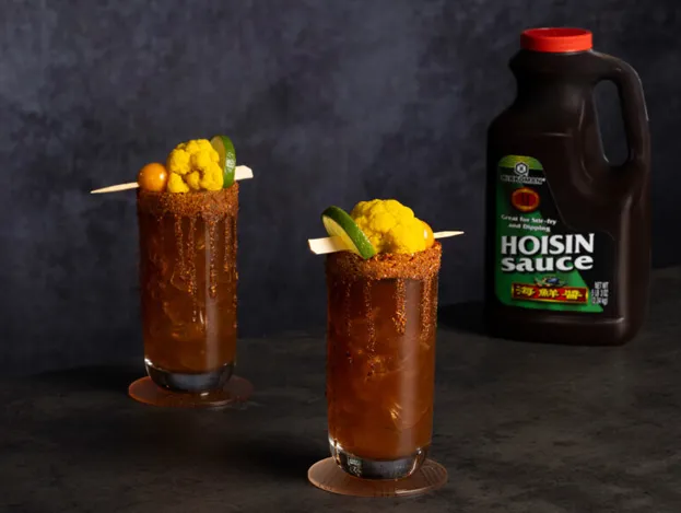 Two glasses of dark-colored cocktails garnished with pickled vegetables and lime, rimmed with seasoning, sit beside a large bottle of hoisin sauce on a dark surface.