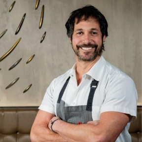 A man with a beard and mustache, wearing a white shirt and a gray apron, stands with arms crossed and smiles in a modern indoor setting.