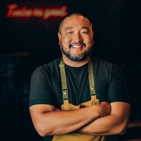 A man with a beard, wearing a black shirt and tan apron, stands with arms crossed, smiling in front of a dark background with red neon writing.
