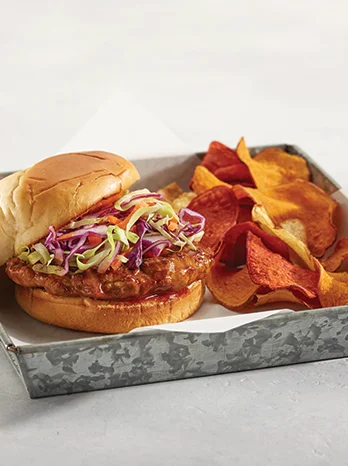 A fried chicken sandwich topped with coleslaw is served in a tray alongside a portion of assorted potato chips.