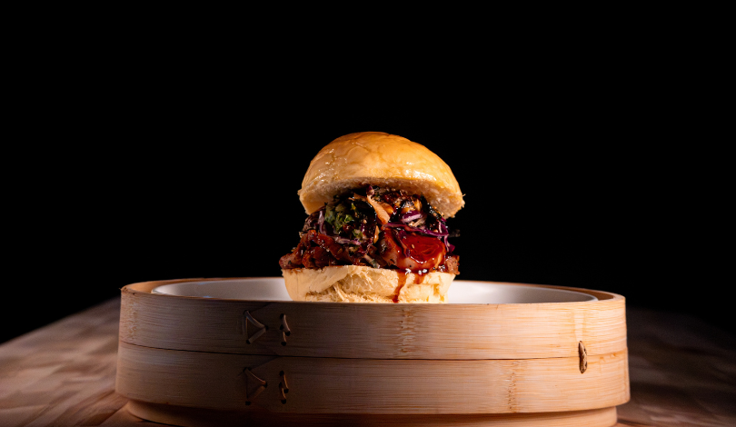 A sandwich with various fillings including vegetables and sauce is placed on a white plate inside a round bamboo steamer, set against a dark background.