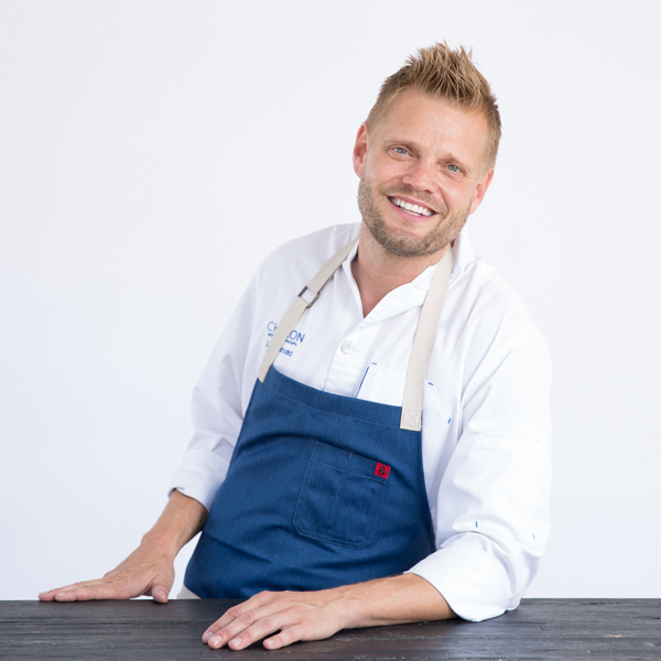 A man wearing a white chef’s coat and blue apron stands behind a black table, smiling at the camera against a plain white background.