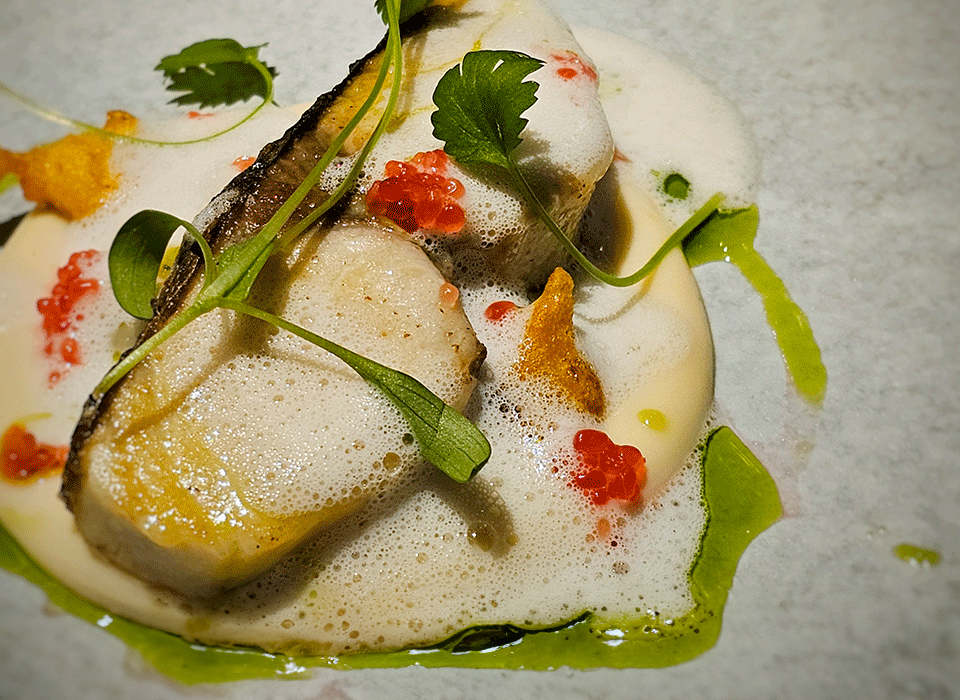 A plated gourmet dish featuring seared fish, foam sauce, red roe, green oil, microgreens, and edible garnishes on a white surface.