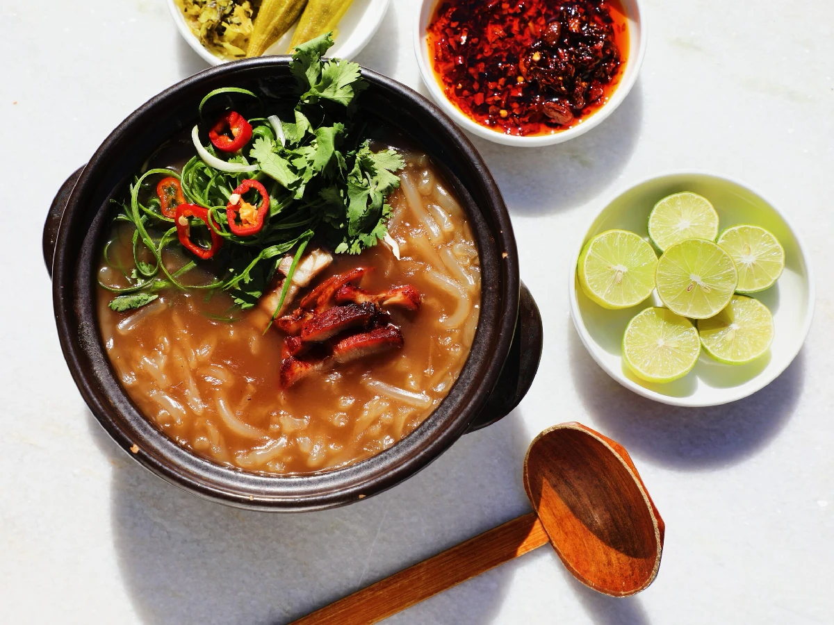 A bowl of noodle soup with sliced meat, herbs, and chili, served with lime wedges, chili oil, pickles, and a wooden ladle on the side.