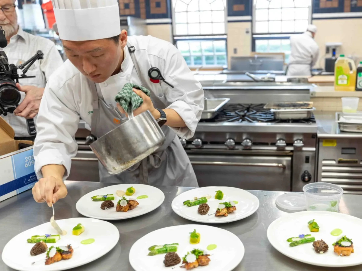 A chef in a white uniform and hat plates food in a professional kitchen, carefully arranging ingredients on several white dishes lined up on a counter.