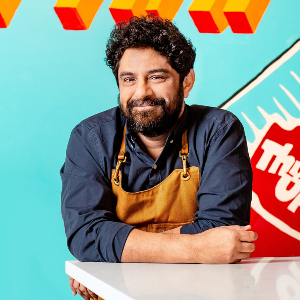 A man with dark hair and beard, wearing a brown apron over a navy shirt, sits at a white counter in front of a colorful background with geometric shapes and text.