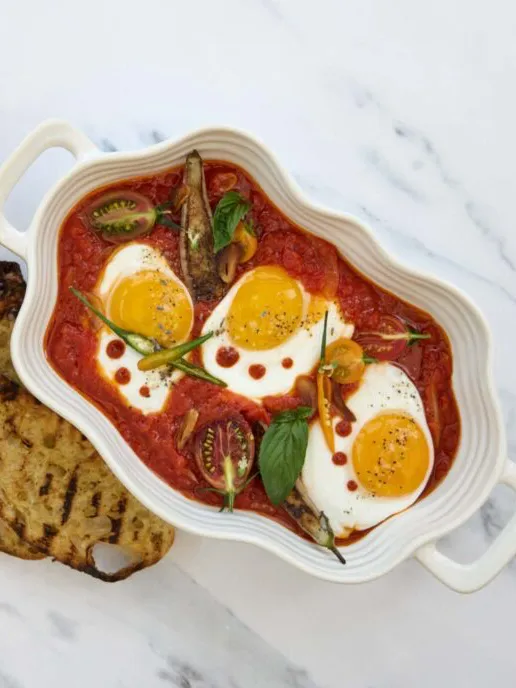 A baked egg dish with three eggs in tomato sauce, garnished with basil, green tomato slices, and served with grilled bread on the side.
