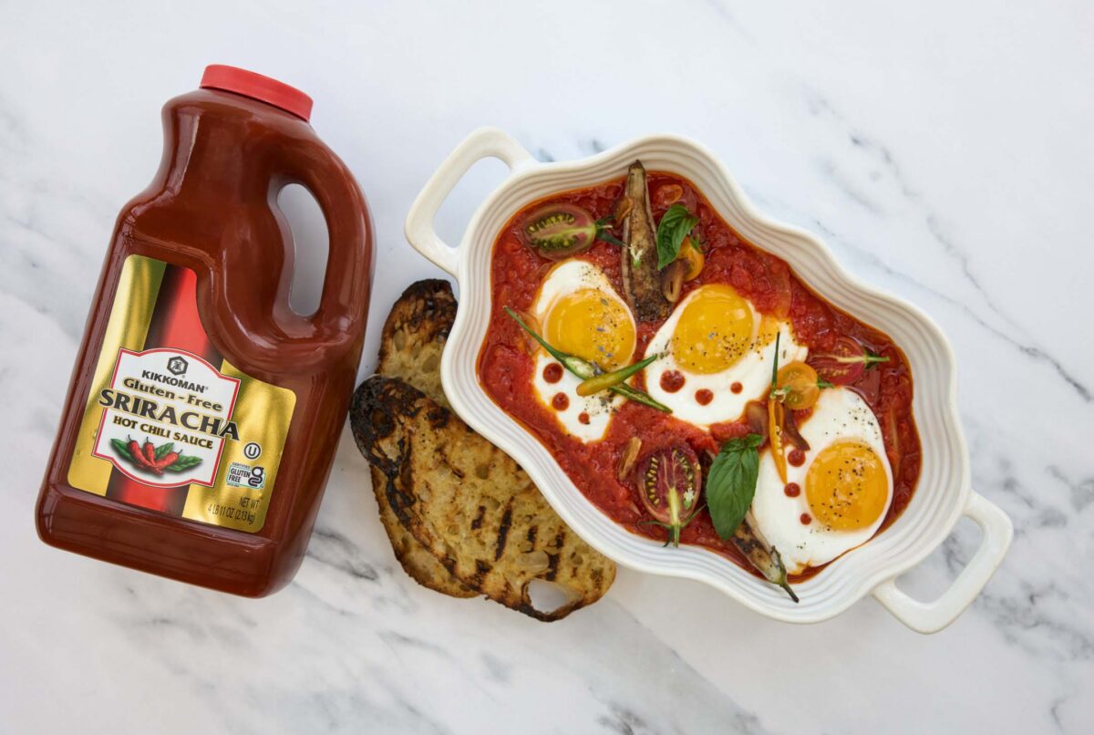 A white dish with three sunny-side-up eggs in tomato sauce, grilled bread slices, and a large bottle of gluten-free sriracha hot chili sauce on a marble surface.