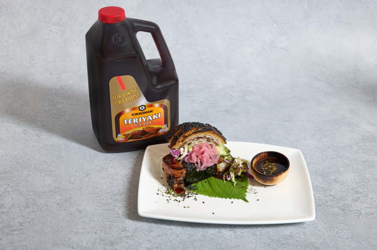 A square white plate with a gourmet dish featuring sliced meat, pickled onions, greens, and sauce, next to a large bottle of Kikkoman Teriyaki sauce on a gray surface.