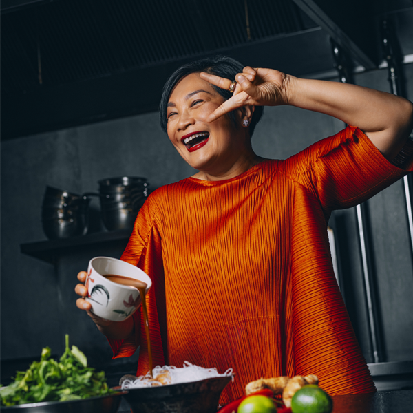 A woman in an orange top smiles and makes a peace sign while holding a cup in a kitchen, surrounded by fresh ingredients on the counter.