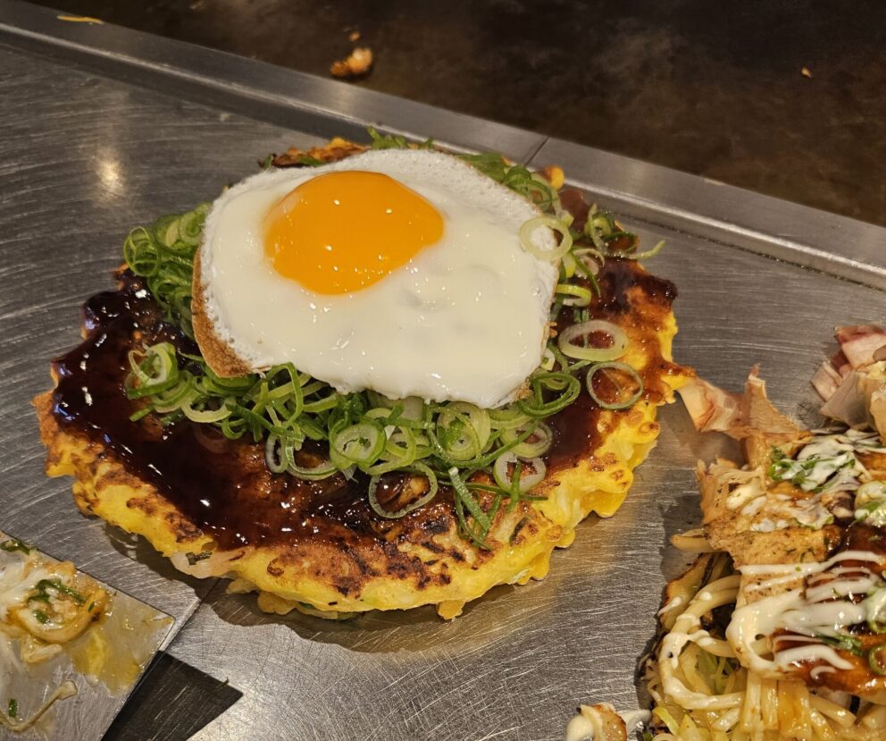 A cooked okonomiyaki pancake topped with a sunny-side-up egg, chopped green onions, and sauce, served on a metal grill.