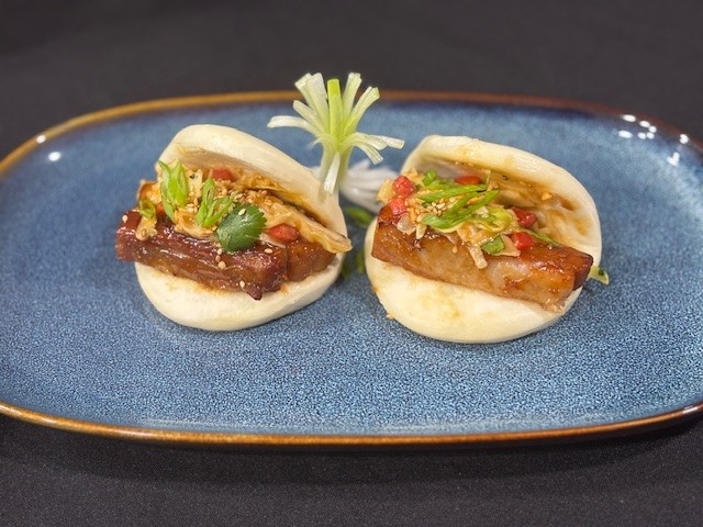 Two steamed bao buns filled with braised pork belly, sliced vegetables, and fresh herbs, served on a blue plate against a black background.