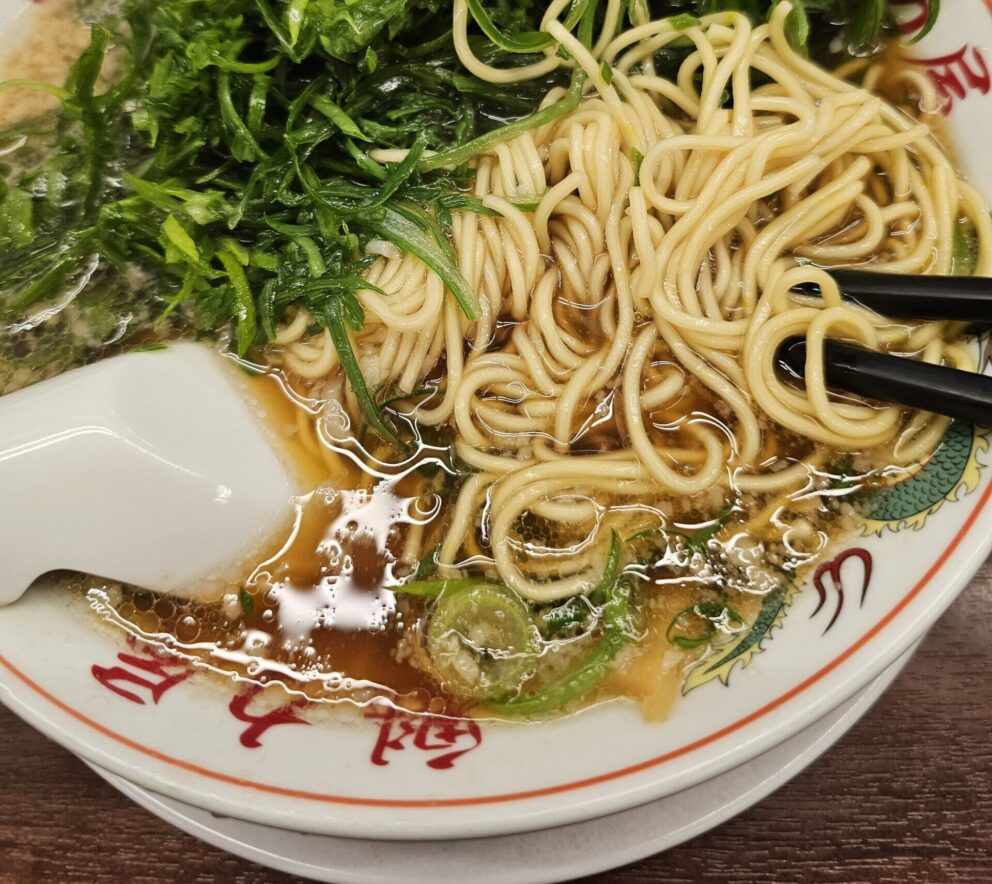 A bowl of noodle soup with green vegetables, broth, and a white ceramic spoon on a decorative plate with red characters.
