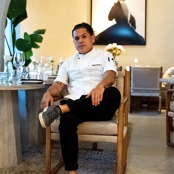 A chef in a white uniform sits on a chair in a modern, elegantly set restaurant dining area with a framed photo in the background.