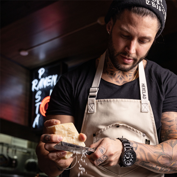 A man wearing an apron and beanie grates cheese over a dish in a restaurant kitchen.