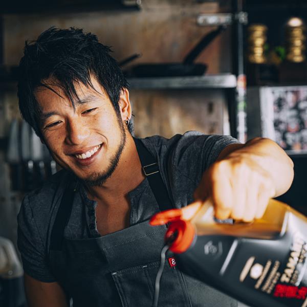 A man wearing an apron smiles while pouring liquid from a large plastic container in a kitchen setting.