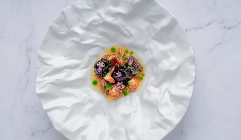 A plated gourmet dish with small portions of meat and vegetables, garnished with herbs and edible flowers, sits on a textured white plate against a marble background.