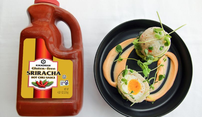 A large bottle of Kikkoman gluten-free sriracha sits next to a black plate with two decorated savory dumplings and sauce swirled on the plate.