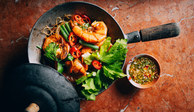 A wok filled with shrimp, noodles, vegetables, and red chili slices, garnished with fresh herbs, next to a small bowl of dipping sauce on a brown surface.