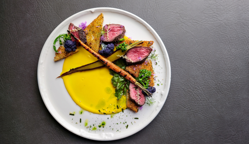 A gourmet dish featuring seared meat slices, toasted bread, carrots, purple vegetables, greens, and yellow sauce, arranged artfully on a round white plate.