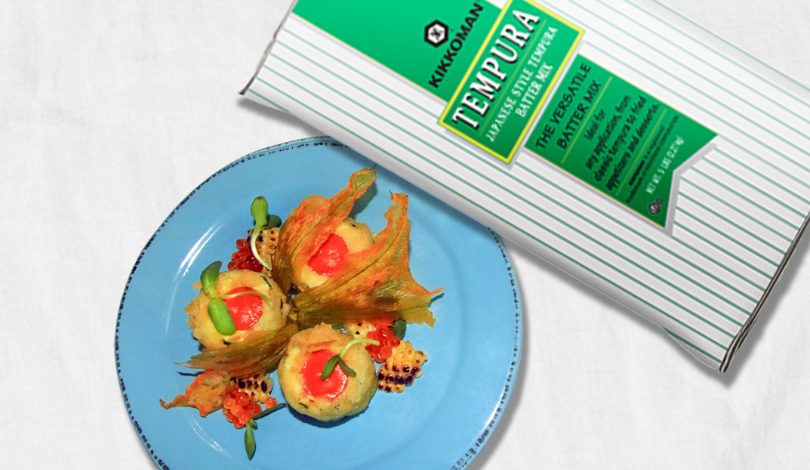 A blue plate with three pieces of vegetable tempura garnished with sauce and greens, next to a box of Kikkoman tempura batter mix.