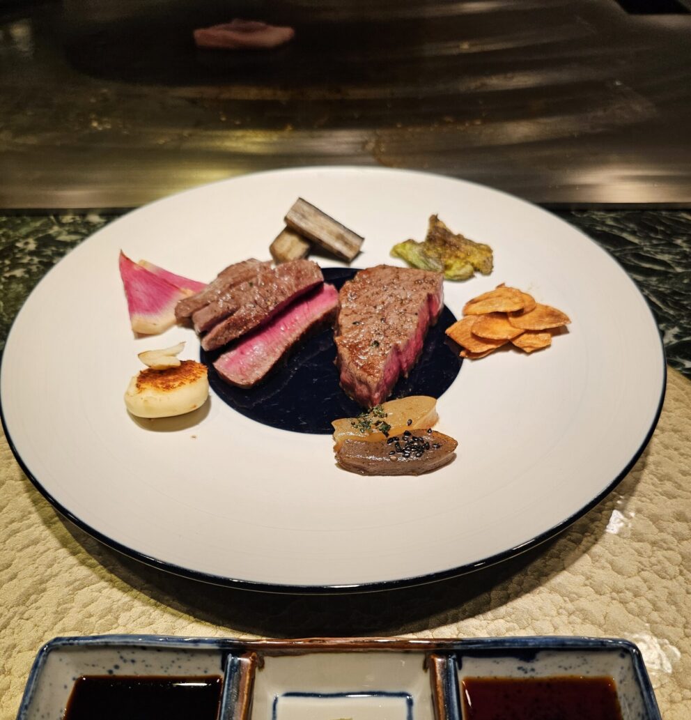 A plated dish featuring slices of medium-rare steak, assorted grilled vegetables, and crisps, with two dipping sauces in small square dishes in the foreground.