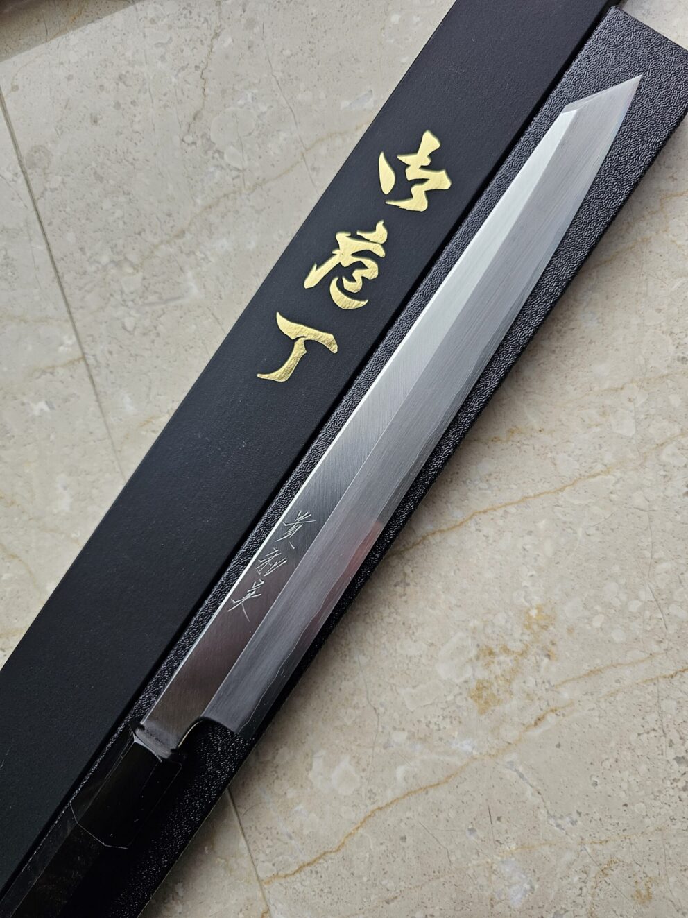 A long, single-edged Japanese knife with a sharp tip is placed next to a black box featuring gold kanji characters on a light-colored stone surface.