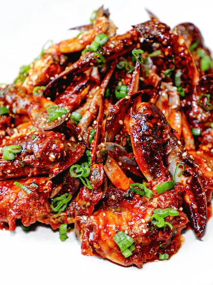 Cooked crab pieces coated in a spicy, dark red sauce, garnished with chopped green onions on a white background.