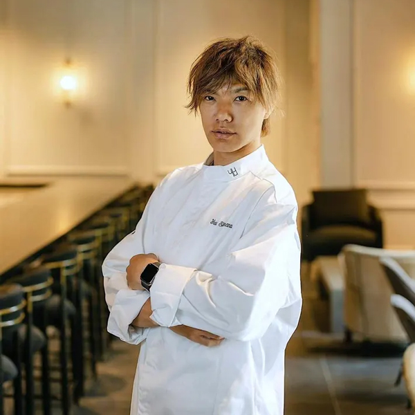 A chef in a white uniform stands with arms crossed in an upscale, modern dining area.