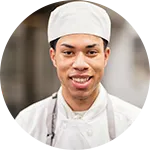 A chef wearing a white uniform and hat smiles at the camera in a kitchen setting.