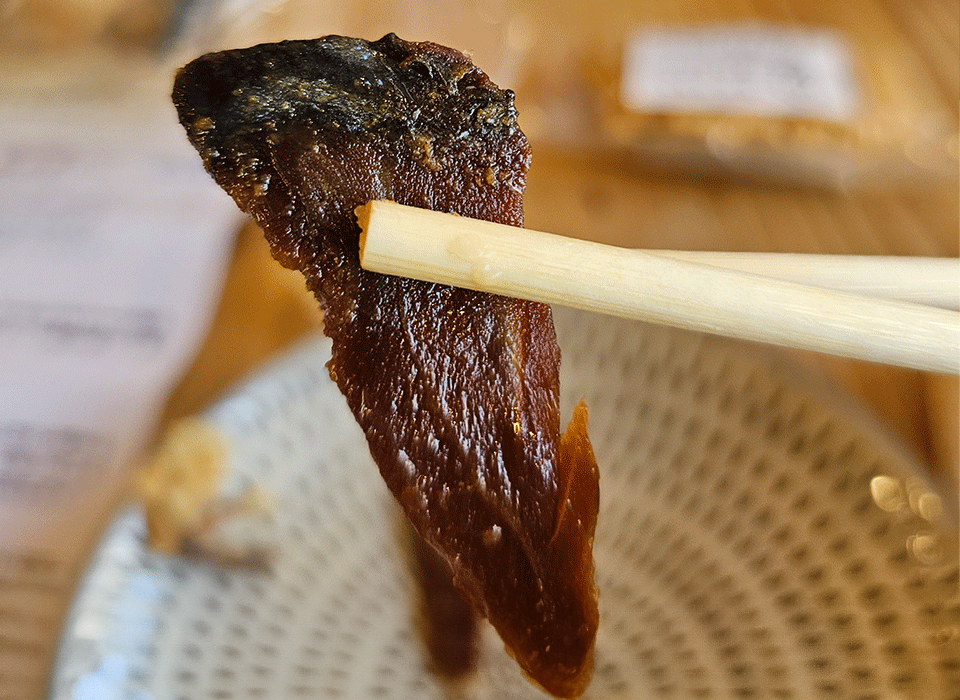 A pair of chopsticks holding a piece of dried fish above a patterned plate.