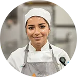 A woman wearing a white chef’s hat and uniform stands in a professional kitchen, looking at the camera and smiling slightly.