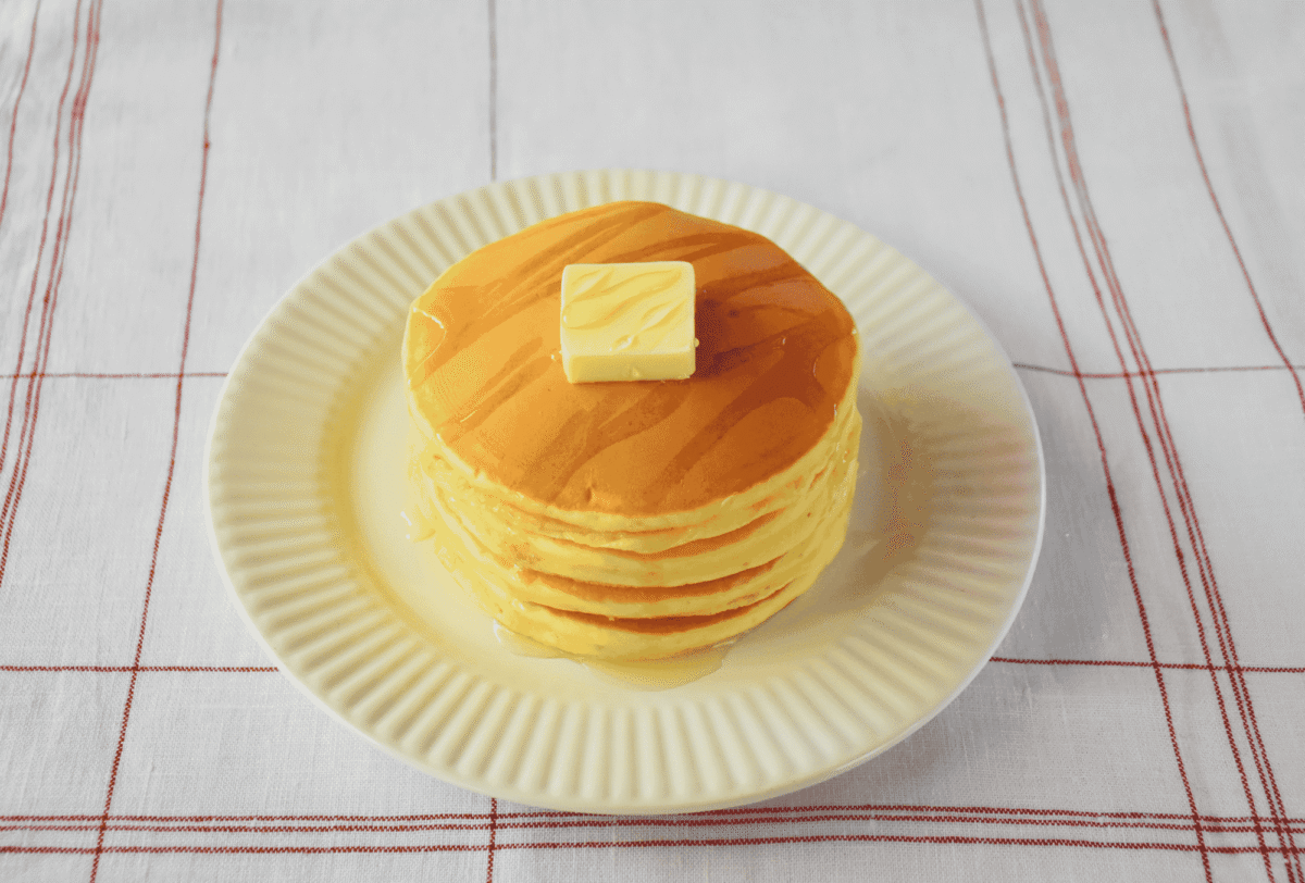 A stack of pancakes topped with a pat of butter and syrup on a white plate, set on a table with a white and red checkered tablecloth.