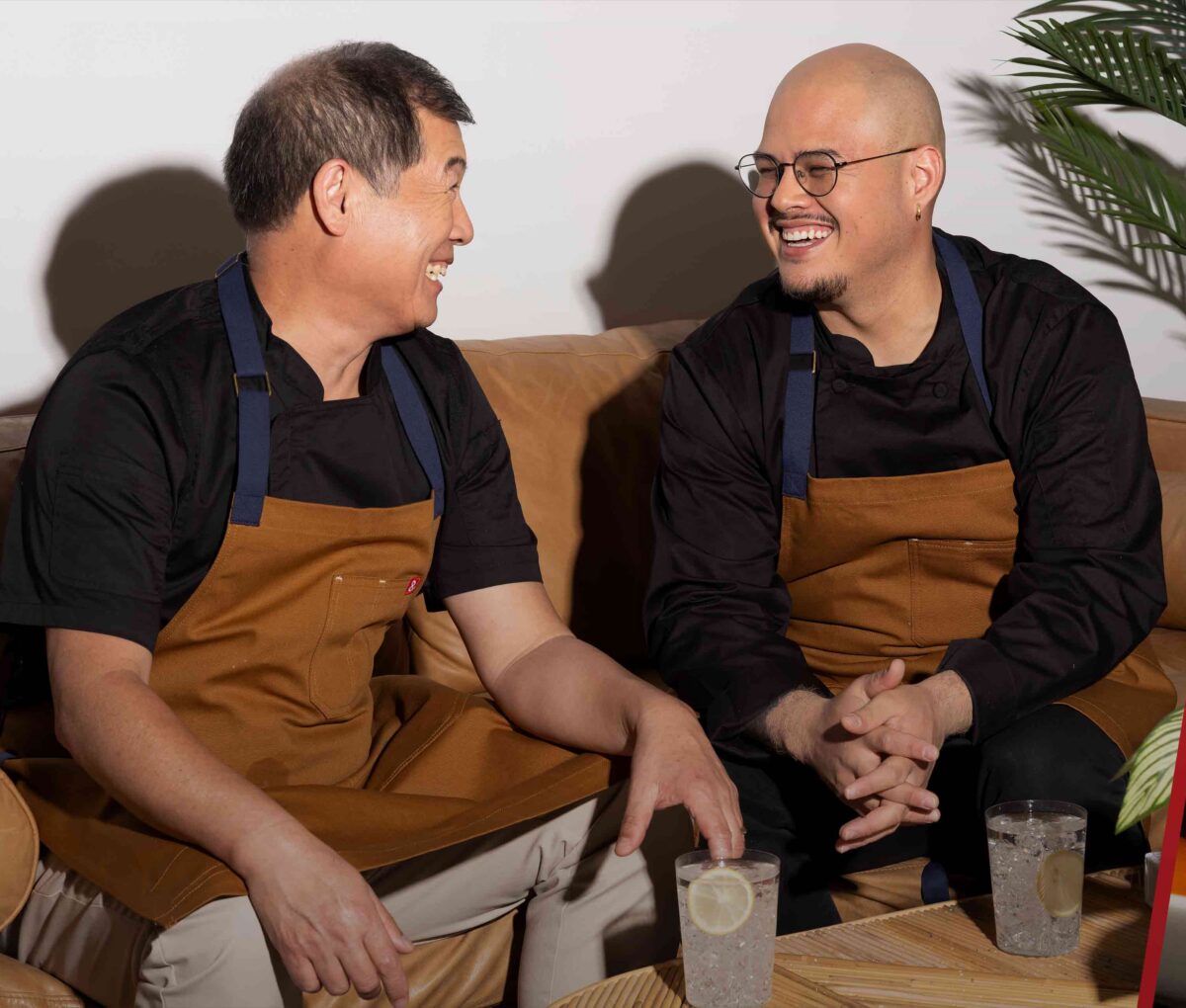 Two chefs sit and talk over drinks between images of plated gourmet dishes, including a black rice dish with dollops and herbs, and an orange soup with white pieces and dark drops.