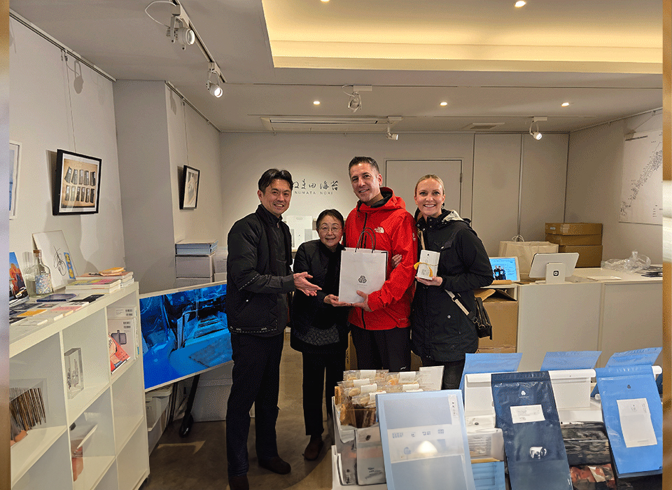 Four people stand smiling inside a small shop with shelves and products displayed. Two of them are holding purchased items. The setting appears modern and well-lit.