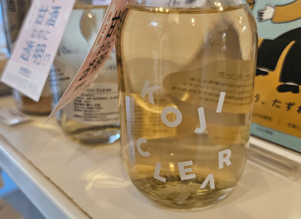 A clear glass bottle labeled "KOJI CLEAR" filled with a light yellow liquid, displayed on a shelf with similar bottles and promotional cards nearby.