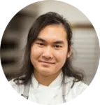A person with long dark hair wearing a white chef's coat looks at the camera and smiles in a kitchen setting.