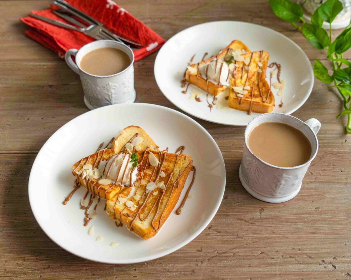Two plates of French toast with cream and syrup, each beside a cup of coffee on a wooden table with a red napkin and green plant in the background.