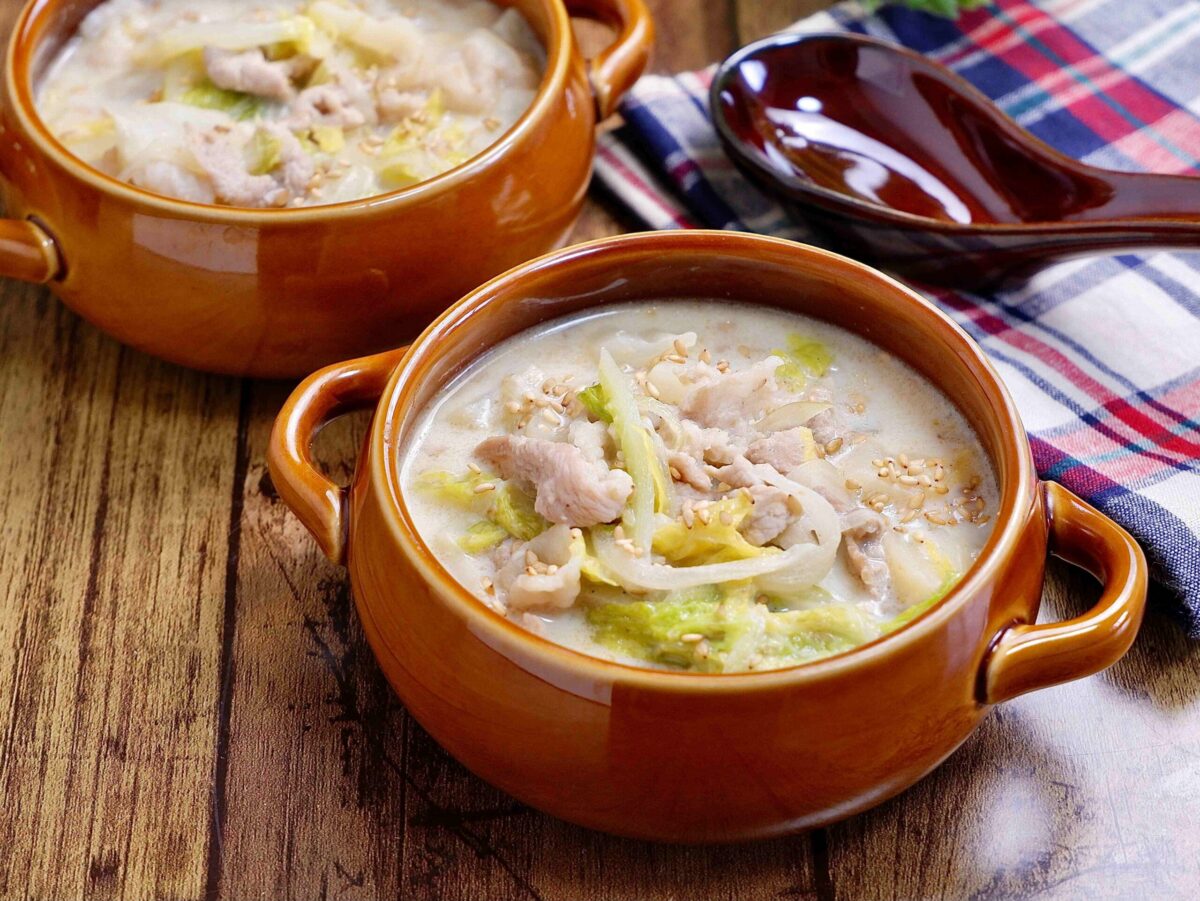Two brown bowls filled with a creamy soup containing sliced meat, cabbage, and sesame seeds, placed on a wooden table next to a plaid cloth and a dark spoon.