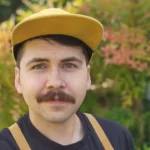 A person with a dark mustache wearing a yellow cap and black shirt with yellow suspenders stands outdoors in front of blurred greenery.