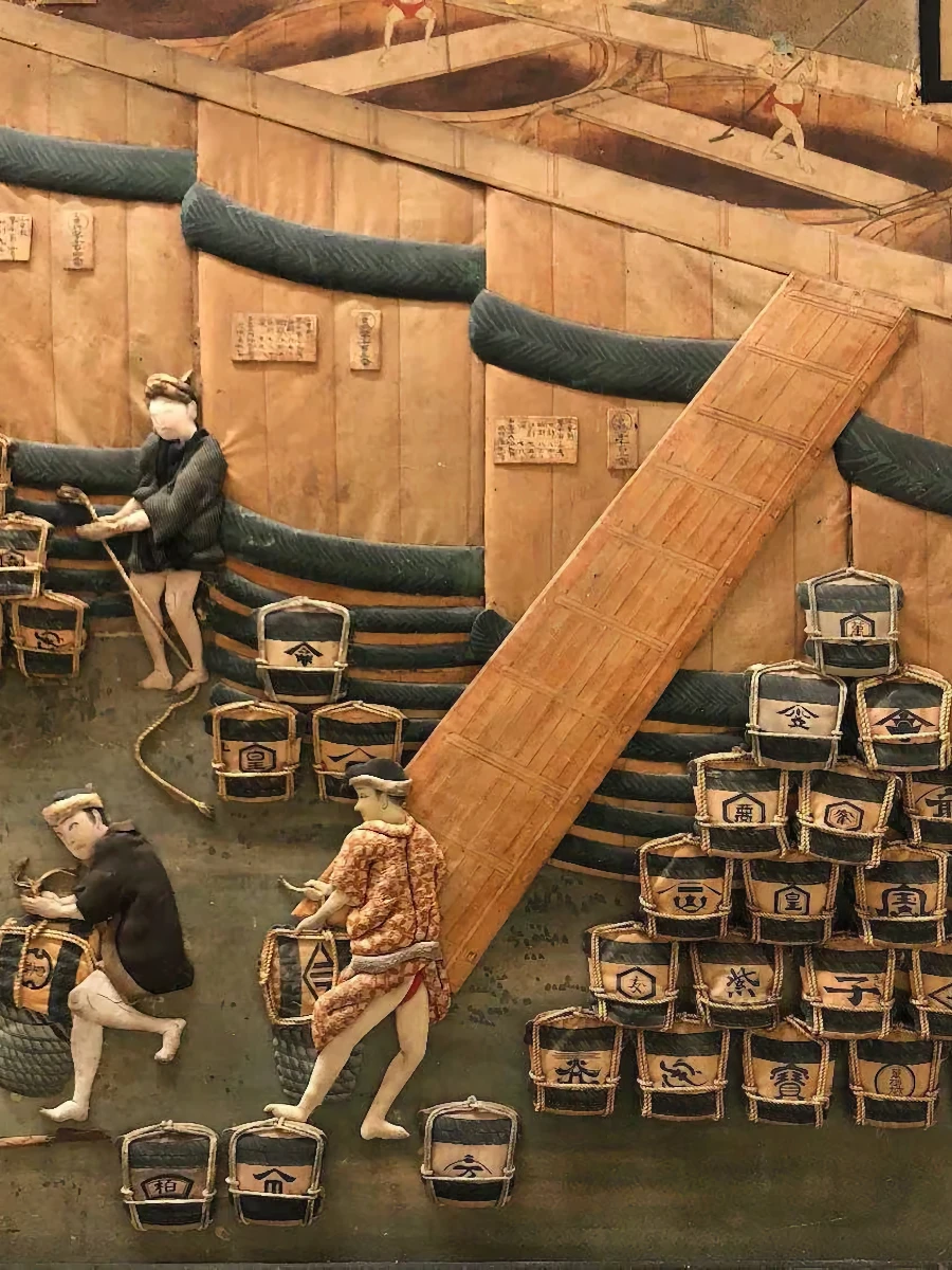Three figures transport sake barrels, stacking them near a wooden ramp, against a backdrop of wooden walls with Japanese signs and labels.