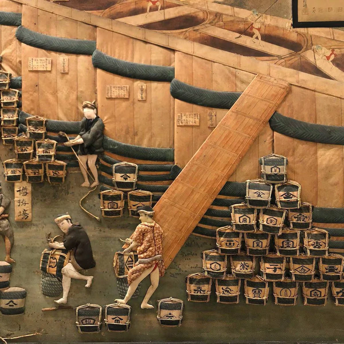 Diorama depicting people transporting and stacking sake barrels with a ramp against a wooden structure, dressed in traditional Japanese attire.