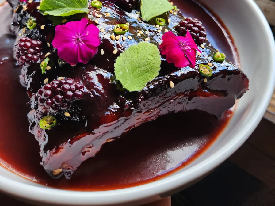 A slab of ribs glazed with a dark sauce, garnished with blackberries, green herbs, pink edible flowers, and sprinkled seeds, served in a white bowl with extra sauce.