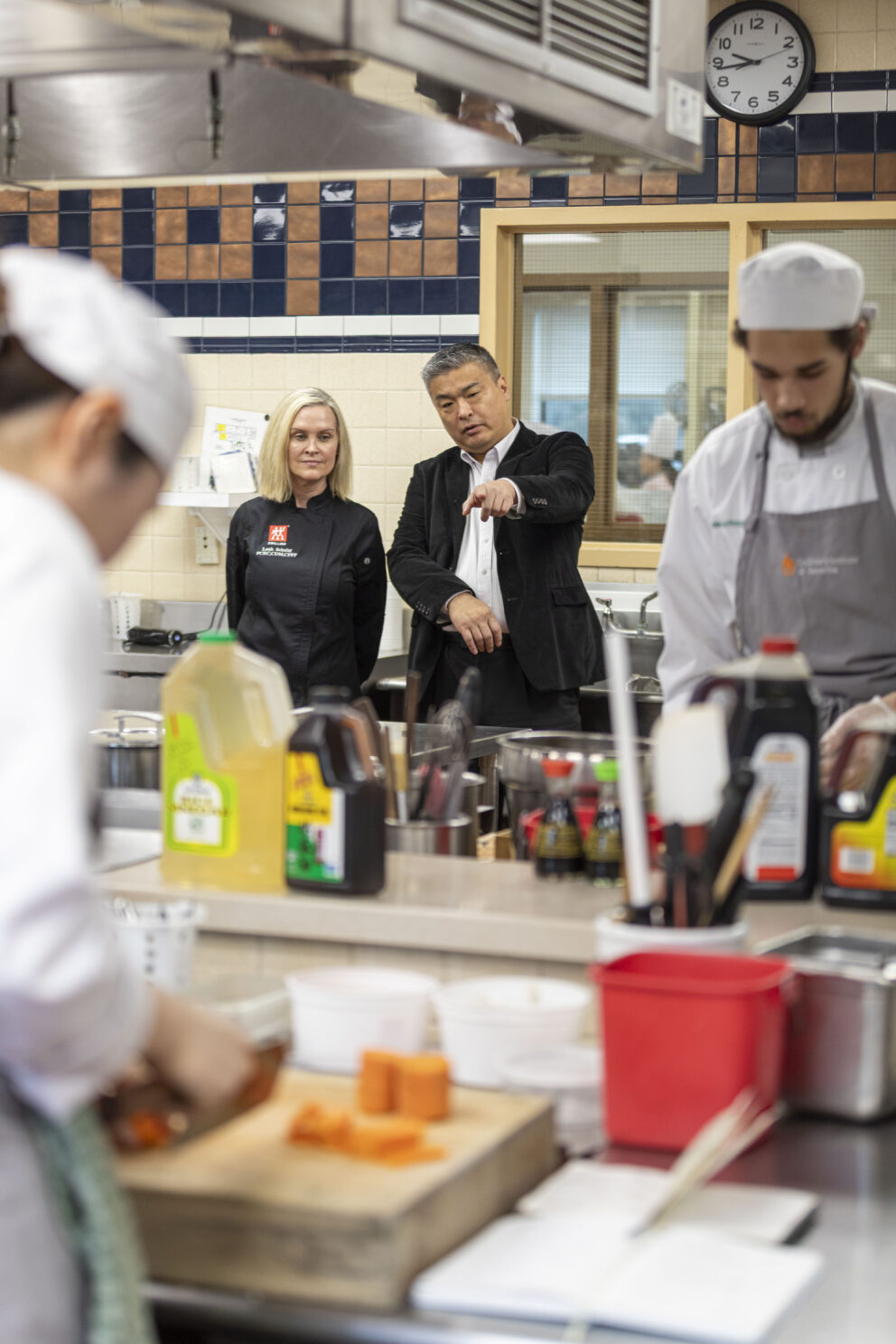Two people observe culinary students preparing food in a commercial kitchen, with various ingredients and cooking equipment visible on the counters.