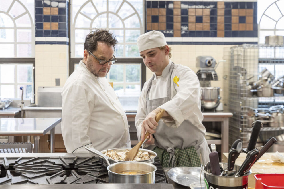 Two chefs in a commercial kitchen; one observes while the other stirs food in a large bowl. Cooking equipment and utensils are visible in the background.