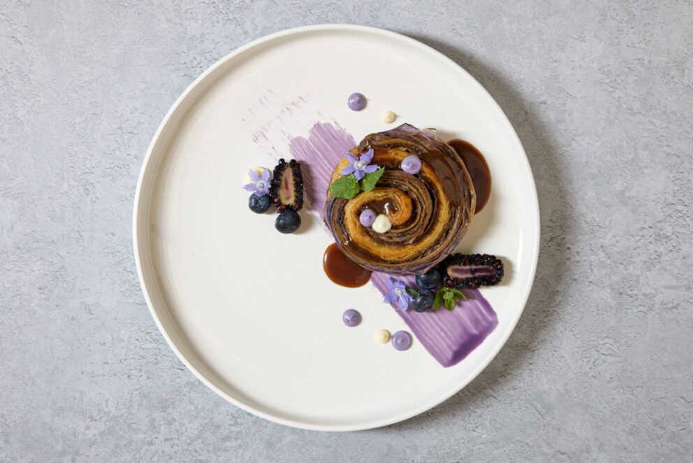 A dessert pastry is plated with blackberry, blueberry, sauce, cream dots, and edible flowers on a white plate with a purple brushstroke, set against a gray background.