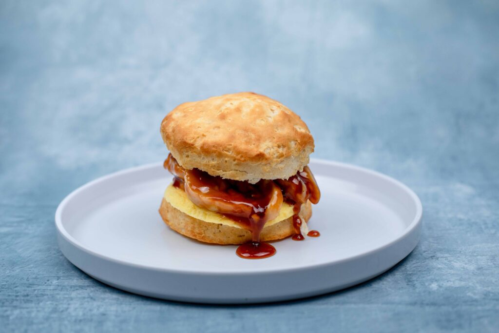 A biscuit sandwich filled with egg, bacon, and drizzled with brown sauce sits on a white plate against a blue background.
