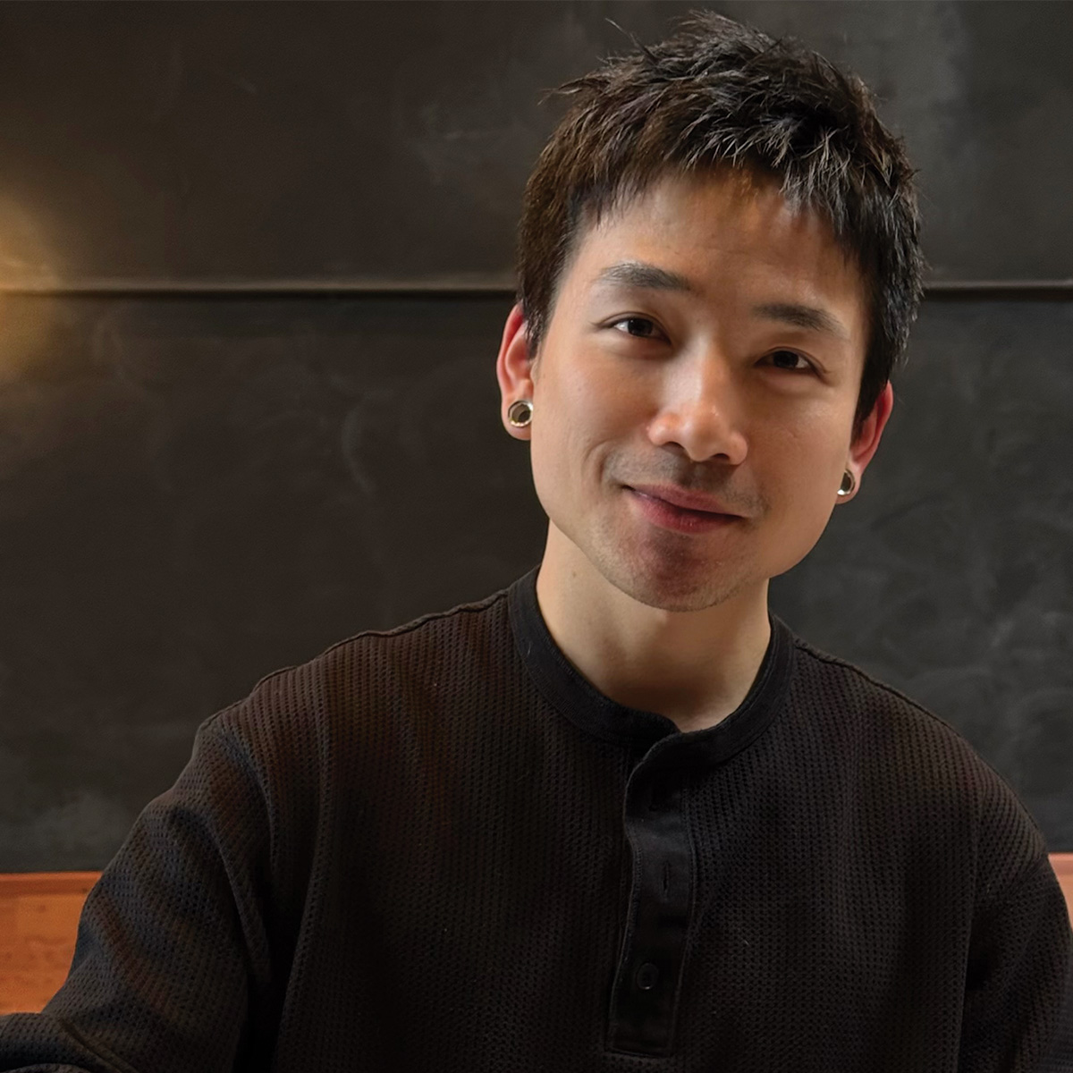 A person with short dark hair, wearing a black textured shirt and ear gauges, sits indoors against a dark background and smiles at the camera.